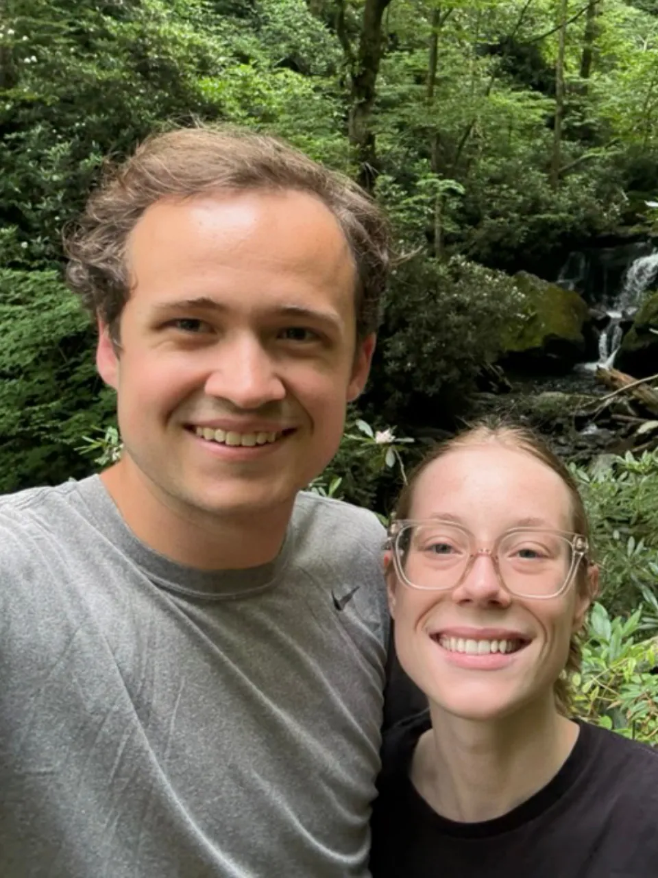 Josh and Jess smiling in a selfie in front of a forest waterfall