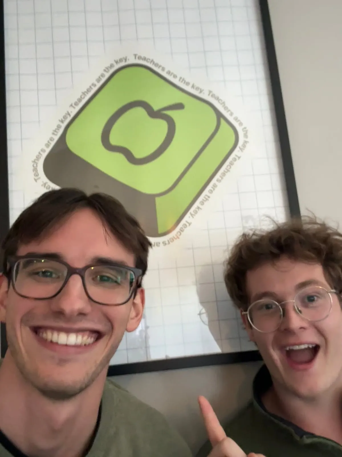 Duncan and Elliot taking a smiling selfie in front of the framed teacher.dev “Teachers are the key” poster